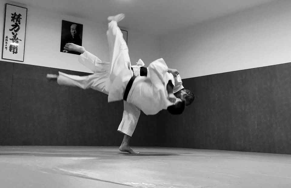 Projection de judo au dojo, sous la calligraphie et le portrait du fondateur